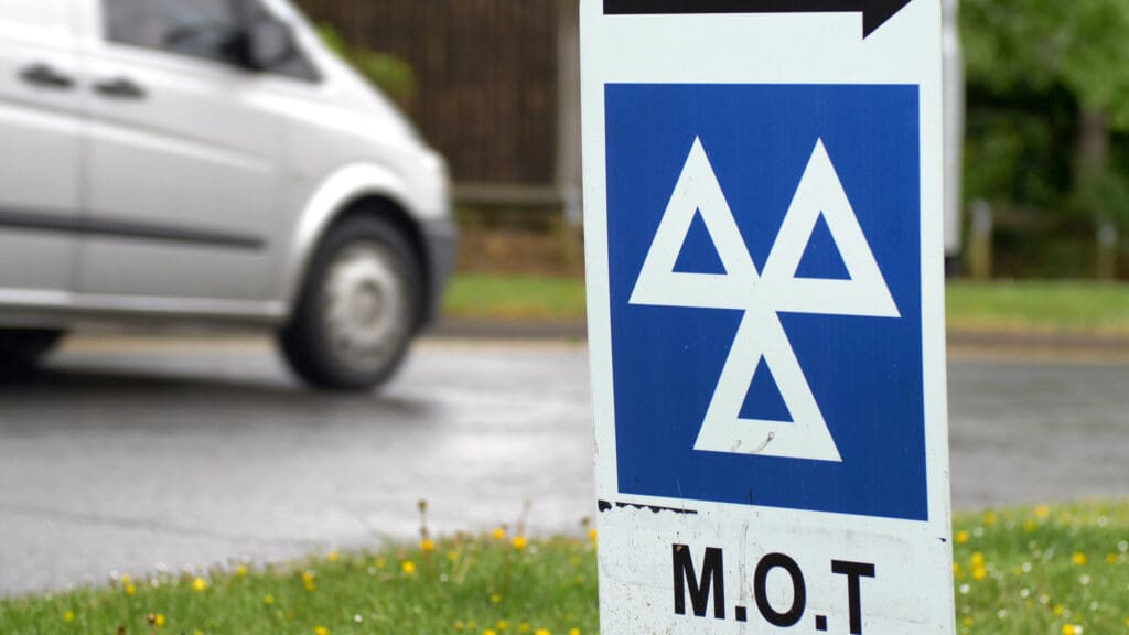 A close-up of an MOT test sign with a blue and white symbol near an Approved Garage. A silver car drives by on a wet road in the blurred background, with grass and small yellow flowers visible near the sign.