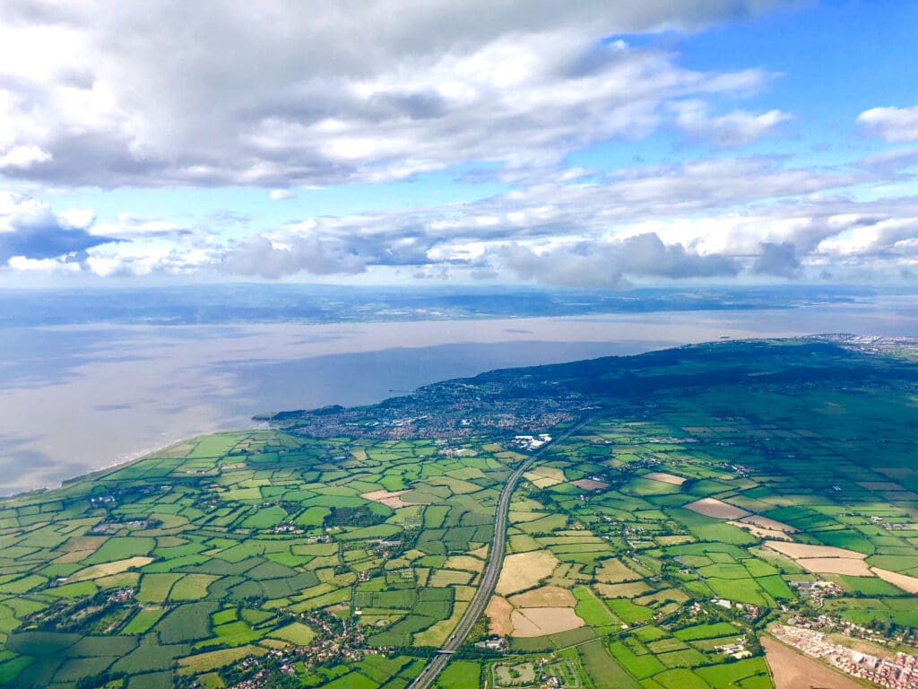 Aerial view of patchwork green and tan farmland, a coastline, and a small town—home to an Approved Garage offering expert Car Servicing—under a partly cloudy blue sky, with a wide body of water in the background.