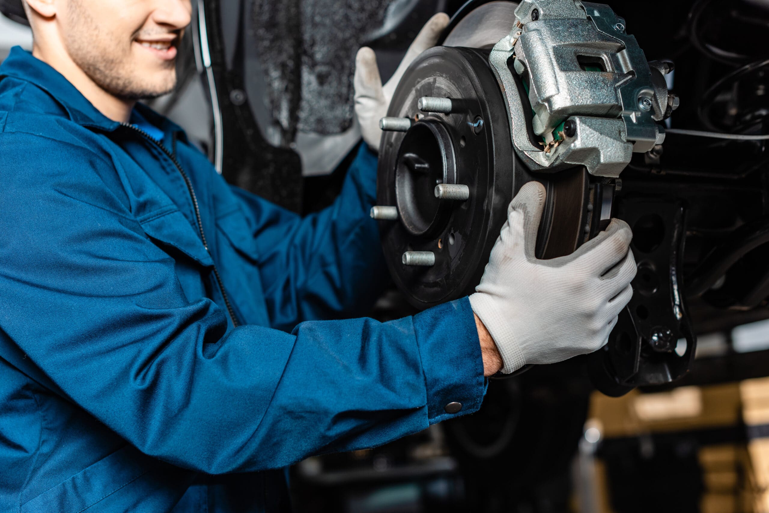 A mechanic in a blue uniform and white gloves inspects a car's brake system, holding the brake disc and caliper assembly during MOT servicing in an approved garage.