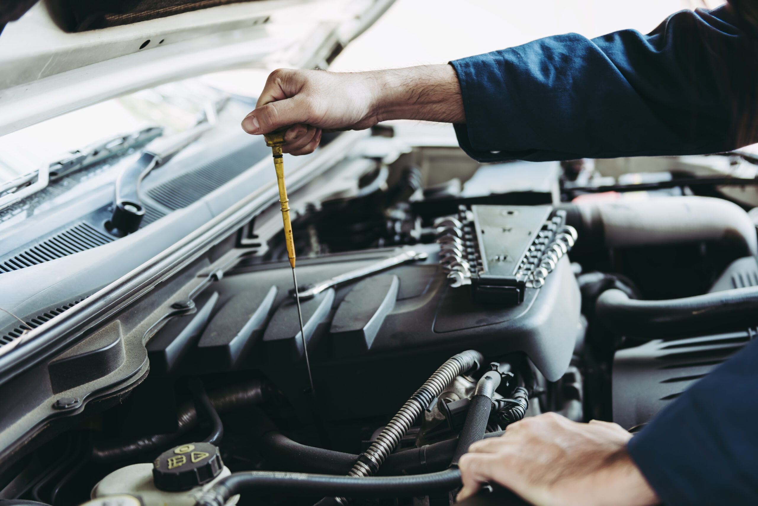 A mechanic in a blue uniform checks the oil level of a car engine by holding and inspecting the dipstick under the open hood at an Approved Garage specializing in MOT Servicing.