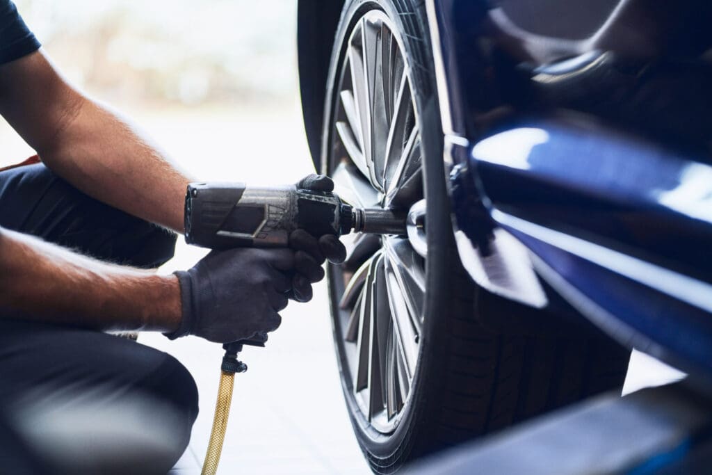 A mechanic wearing gloves uses a power tool to tighten or loosen the lug nuts on a car wheel during professional car servicing at an approved garage.