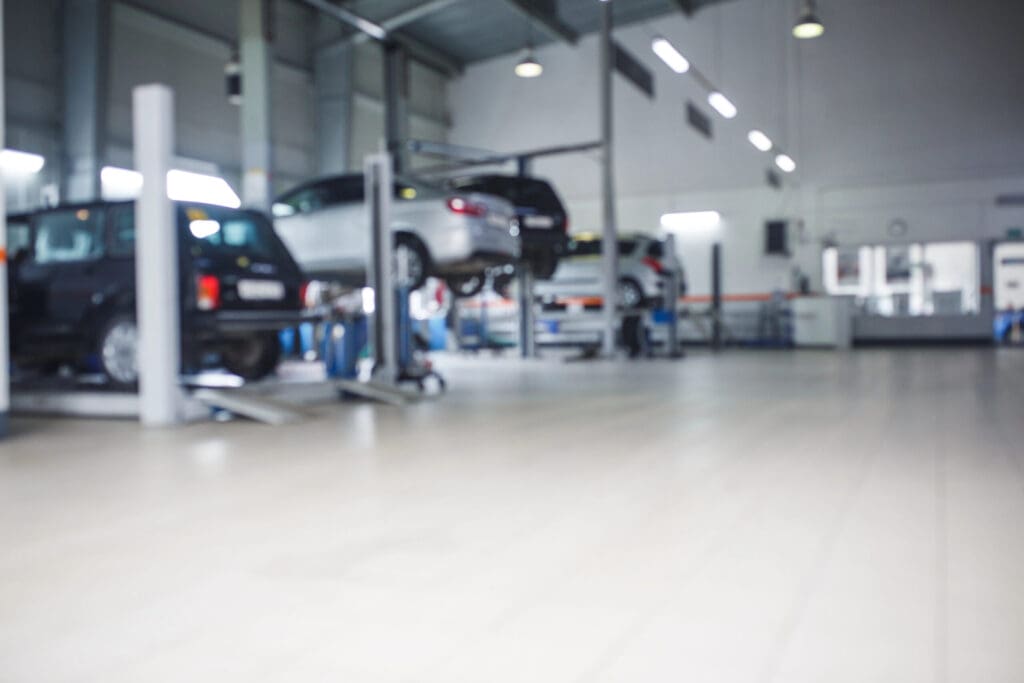 A blurred image of an Approved Garage car repair shop with several cars elevated on lifts, mechanics working, and an open, well-lit space ideal for MOT Servicing—with tiled floors and high ceilings.