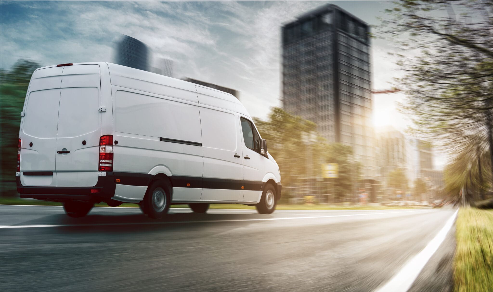 A white delivery van drives quickly down a city street, with motion blur indicating speed. Modern buildings and trees line the road under a bright, sunny sky.