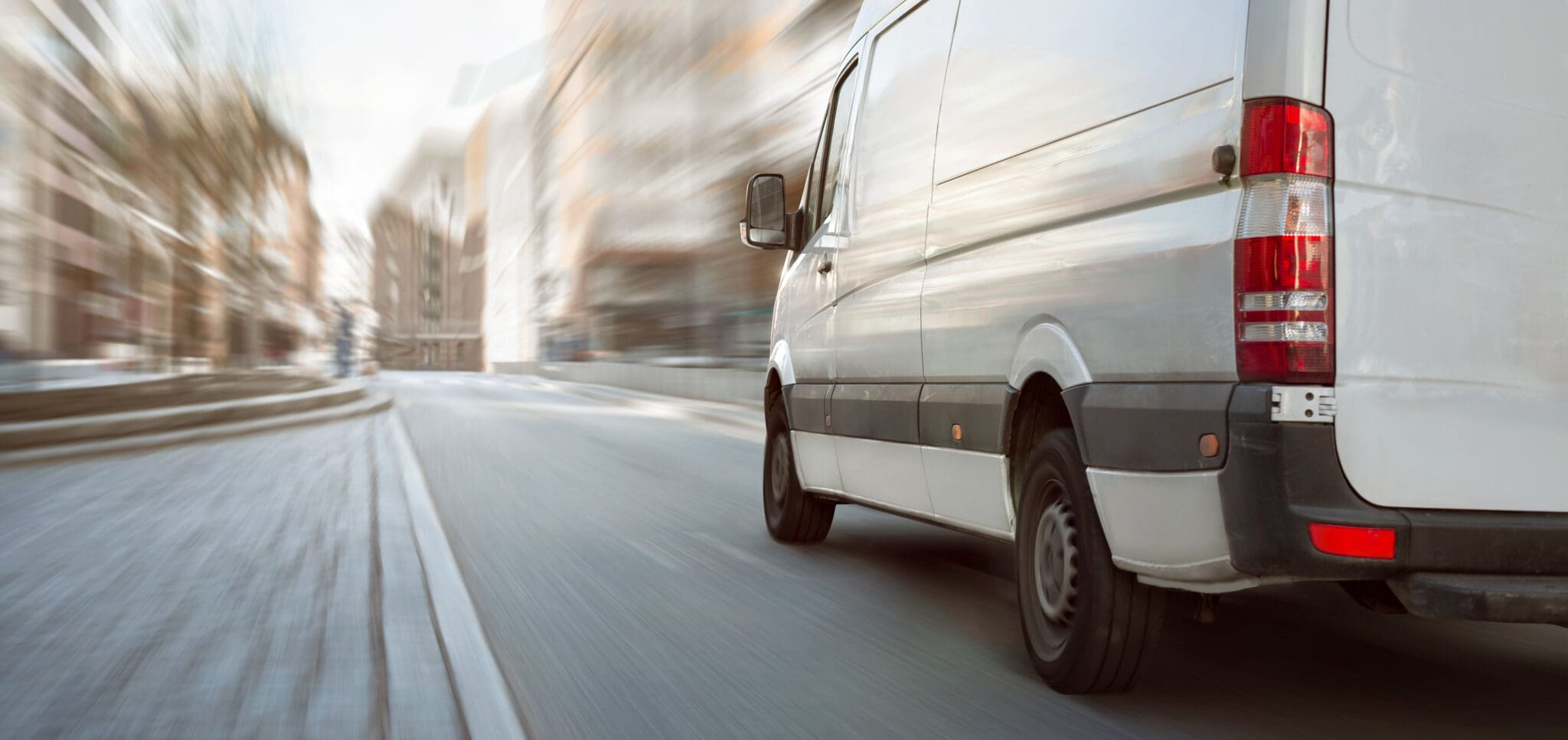 A white delivery van drives quickly on a city street, with buildings blurred in the background to show motion.